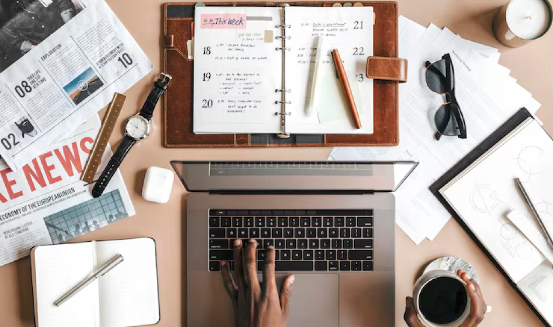 Workspace with laptop, planner, newspapers, and notebooks used for organizing Case Studies & Press Releases, with a person typing and holding a cup of coffee.