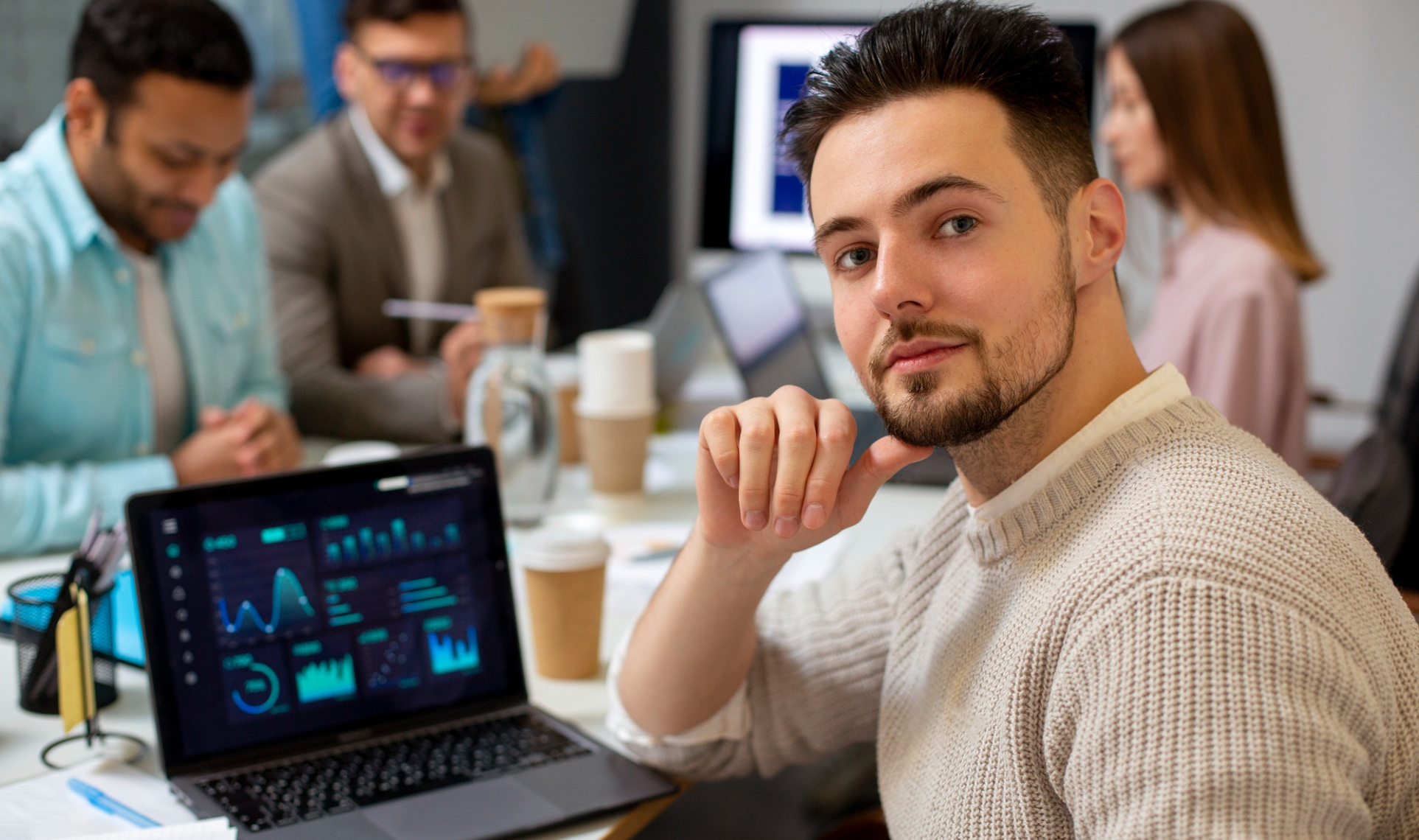 : A focused man in a meeting checking a laptop displaying financial data dashboards, with colleagues in the background.